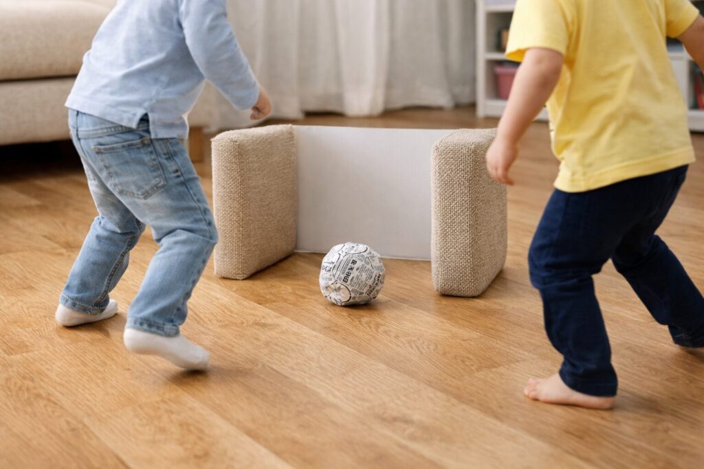 リビングの床で、新聞紙を丸めて作ったボールを使って子どもたちが遊ぶ室内サッカーの風景。クッションと箱を使った簡単なゴールが置かれ、おうちで安全に体を動かして遊べる様子が伝わる画像。