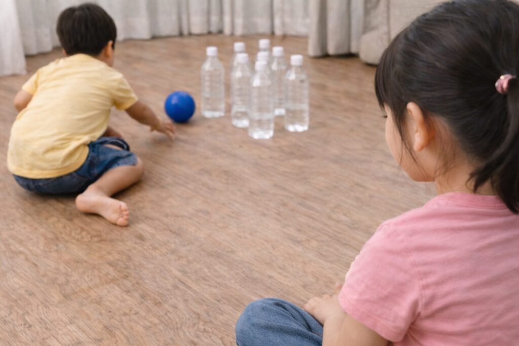 リビングの床に並べた空のペットボトルをピンに見立て、子どもが青いボールを転がして遊ぶペットボトルボウリングの風景。もう一人の子どもが近くで見守っており、おうちで楽しむ室内遊びの様子が伝わるリアルな画像。